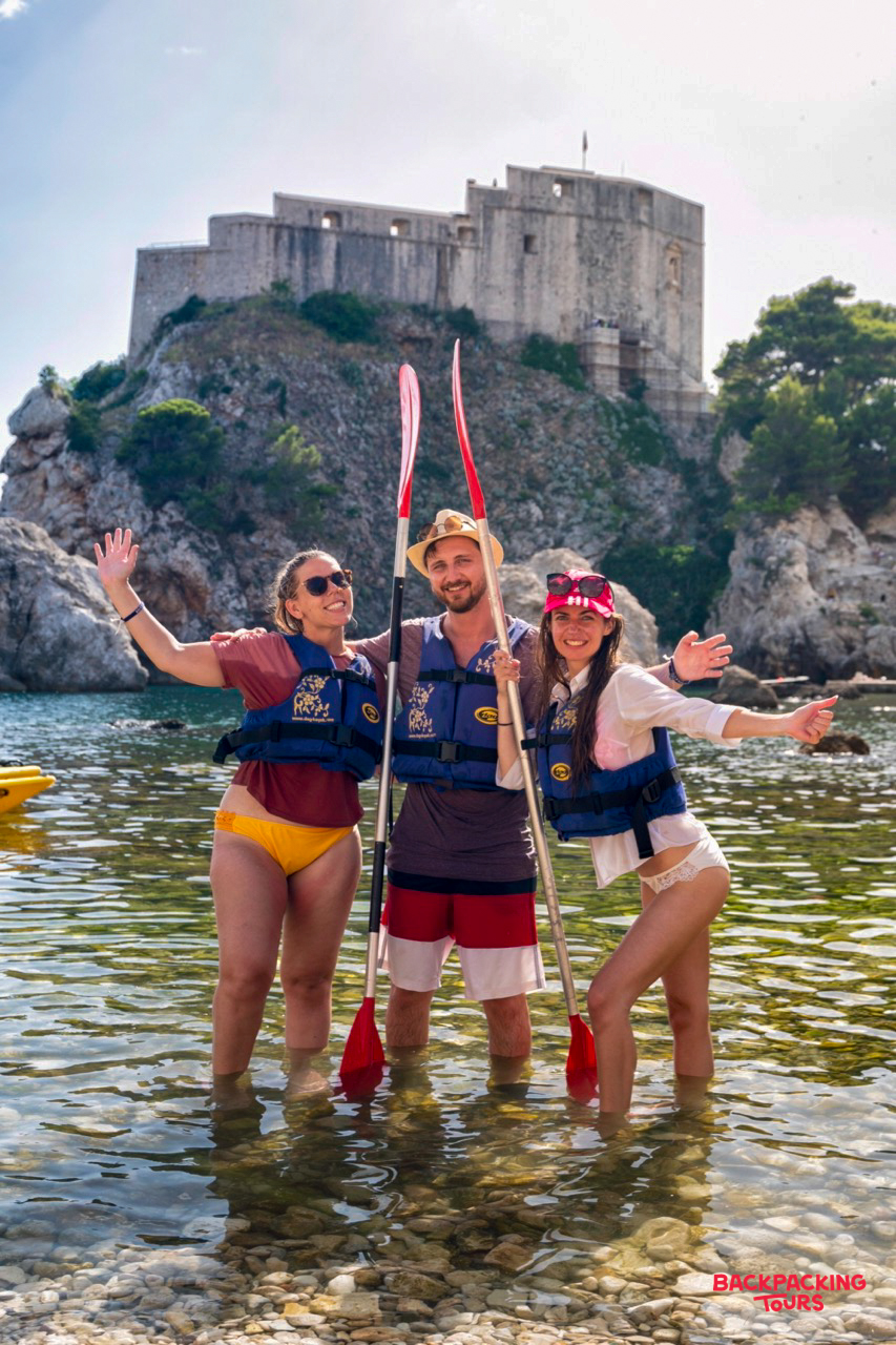 Three happy travelers with kayak paddles stand in the water near Dubrovnik's historic fortress.