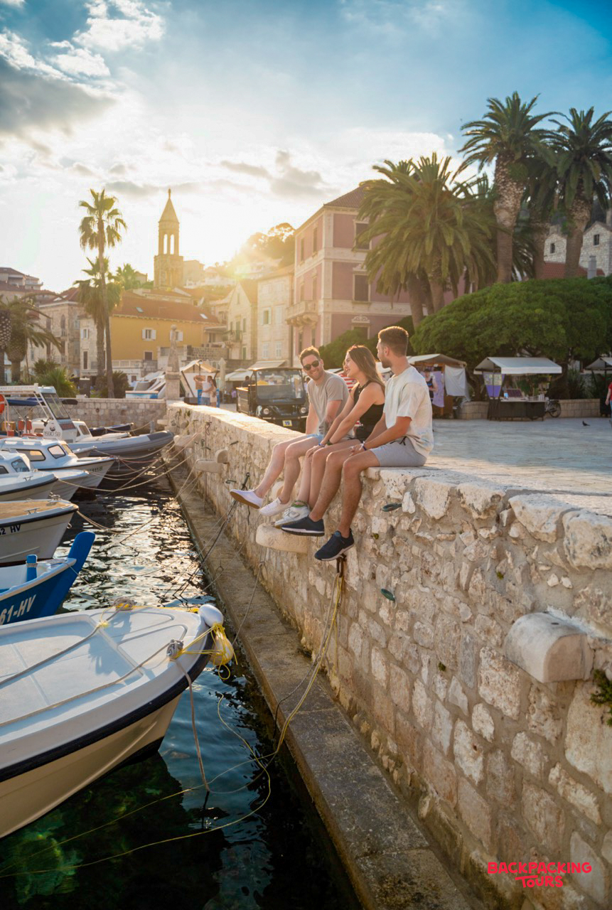 Three friends relax on a stone wall overlooking boats in Hvar harbor during a beautiful sunset.