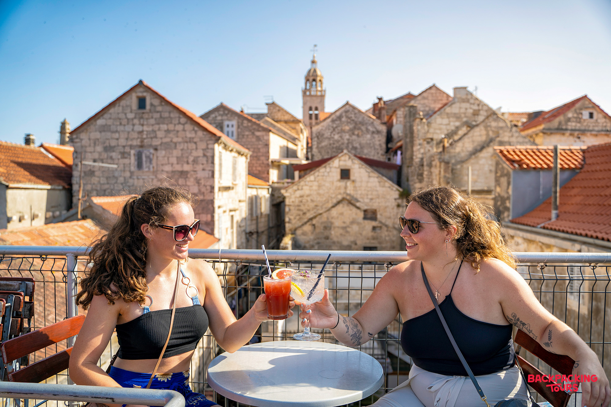Two women toast cocktails on a sunny rooftop terrace overlooking Korcula's old town.