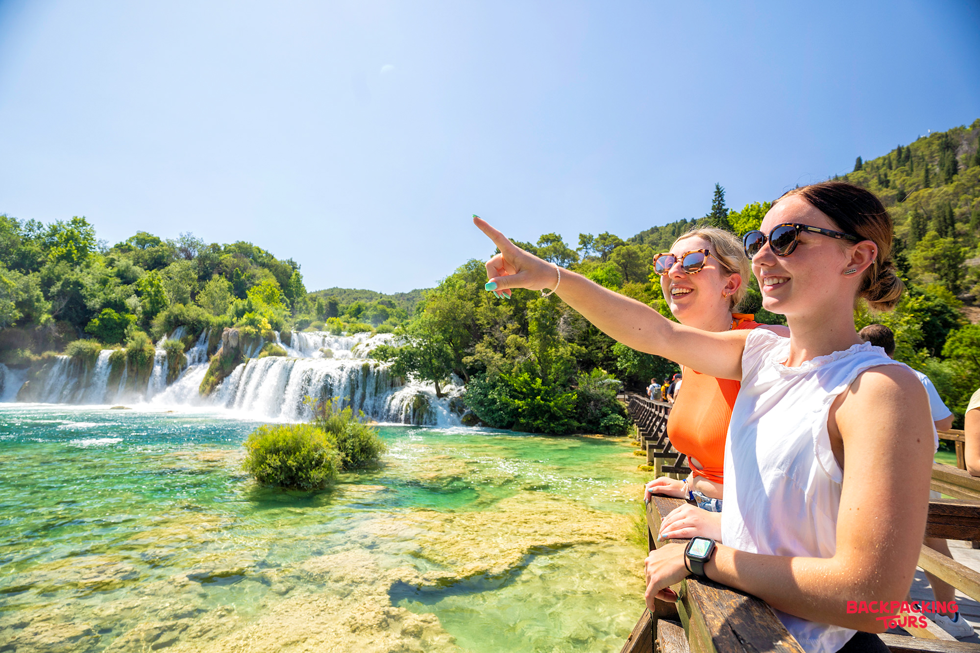 Two happy women pointing at the stunning turquoise waterfalls in Croatia's Krka National Park.