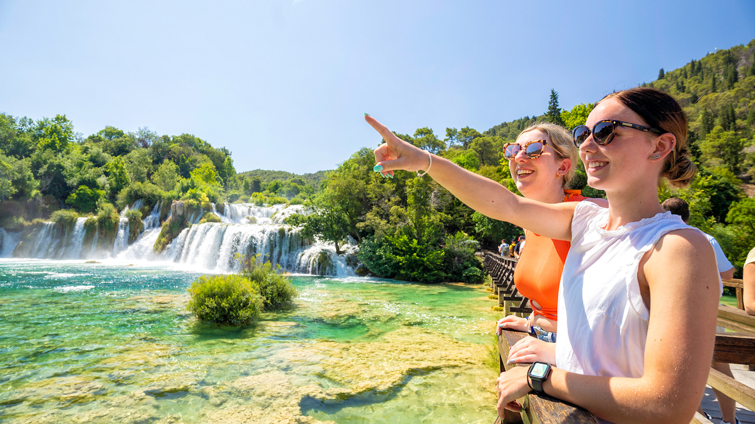 Two smiling women on a boardwalk pointing at the stunning waterfalls in Krka National Park.