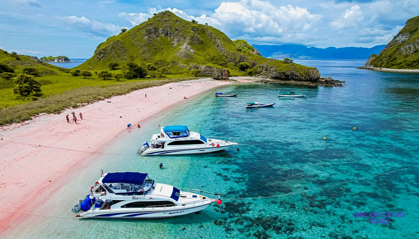 Boats moored in turquoise water off Pink Beach in Komodo National Park, with green hills rising behind.