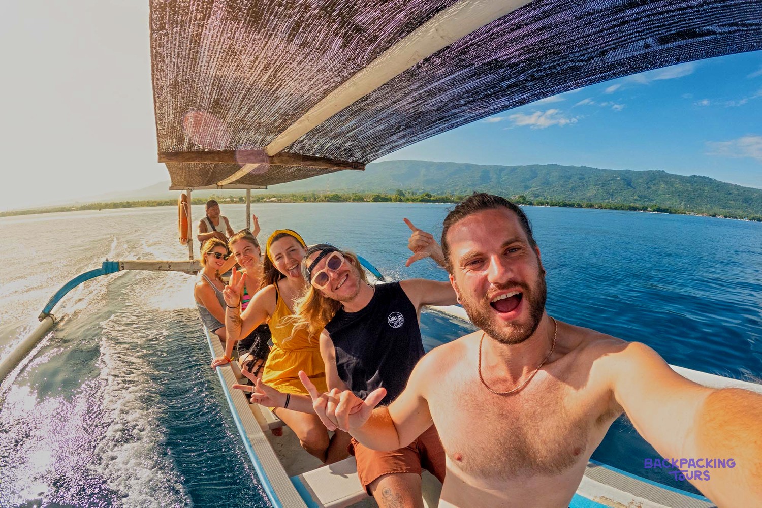 Six smiling people on an outrigger boat on bright blue water with distant hills.