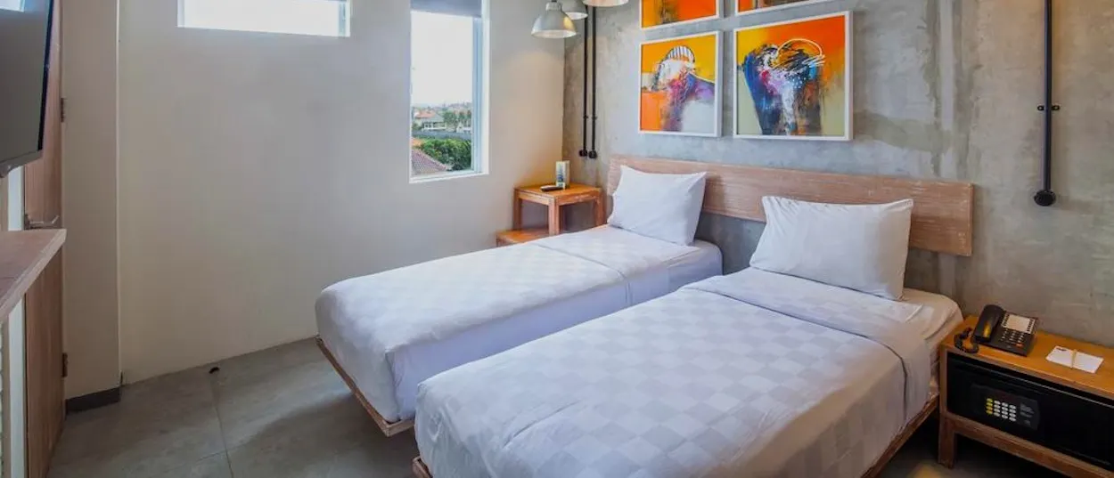 A bright hotel room contains two single beds, white bedding, and colourful abstract artwork on the wall.