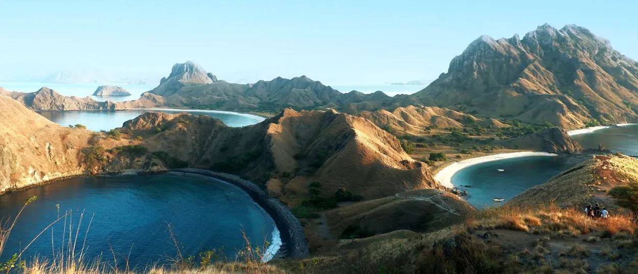 Rolling dry hills surround deep blue bays with white and black sand beaches under a clear sky.