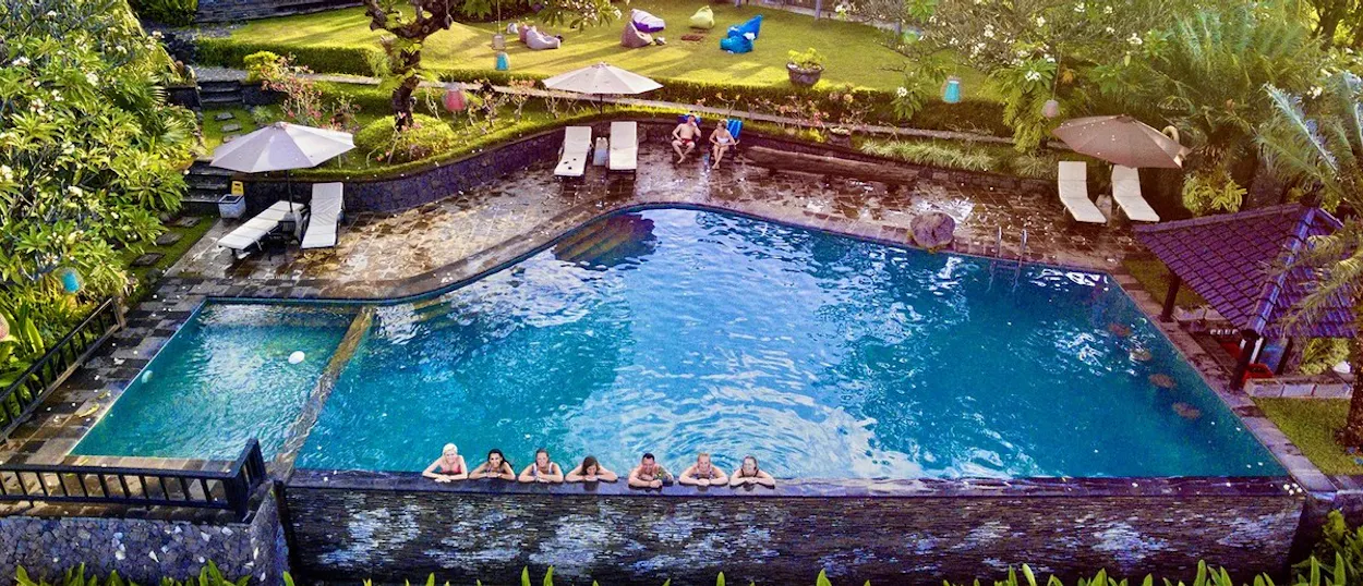 A private villa with a large swimming pool, people relaxing in the water, surrounded by lush gardens and palm trees.