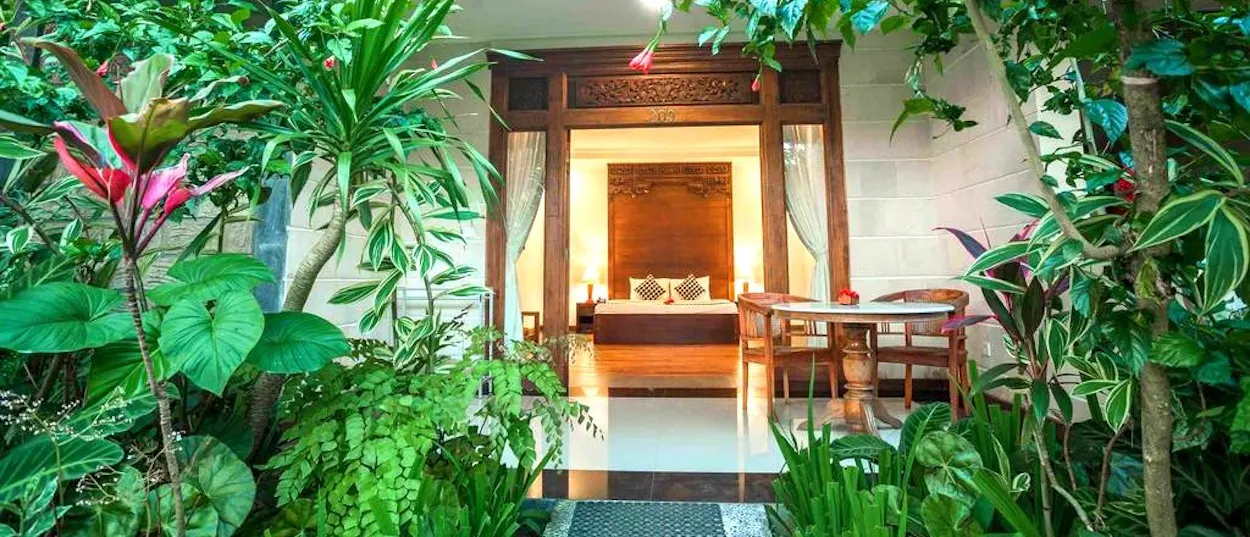 A wooden doorway leads to a bright bedroom with a bed, surrounded by lush tropical foliage.