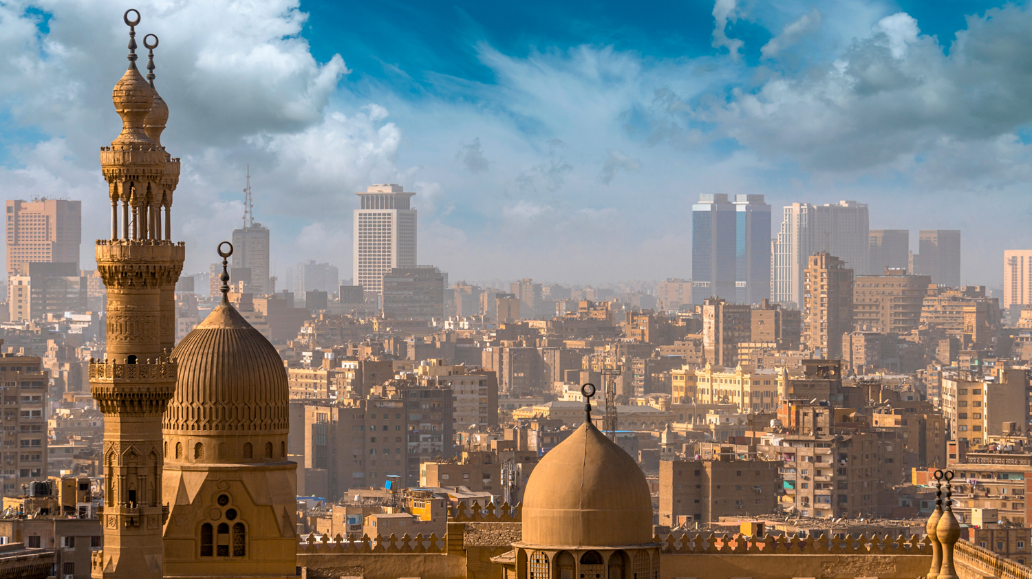 Historic minarets and domes rise above the sprawling city skyline of Cairo under a blue sky.