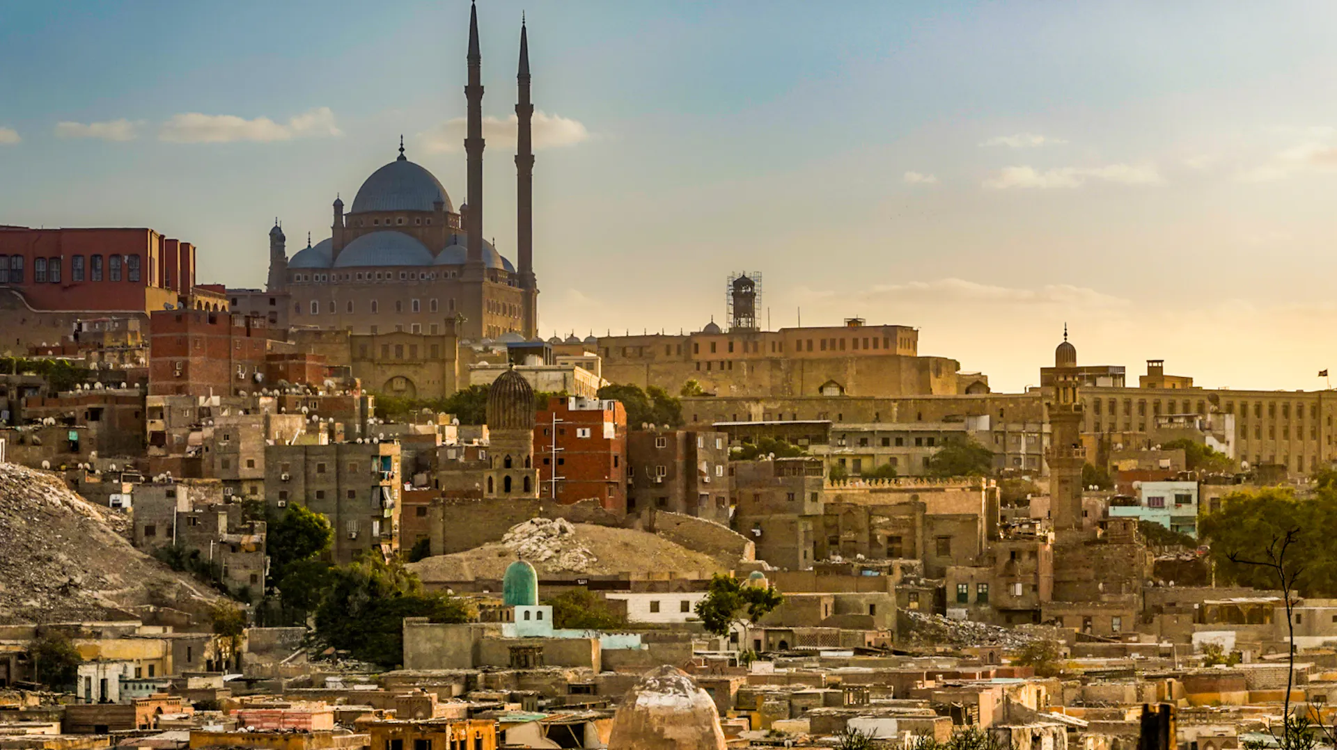 Warm sunset light illuminates the historic cityscape of Cairo, featuring the iconic Muhammad Ali Mosque.
