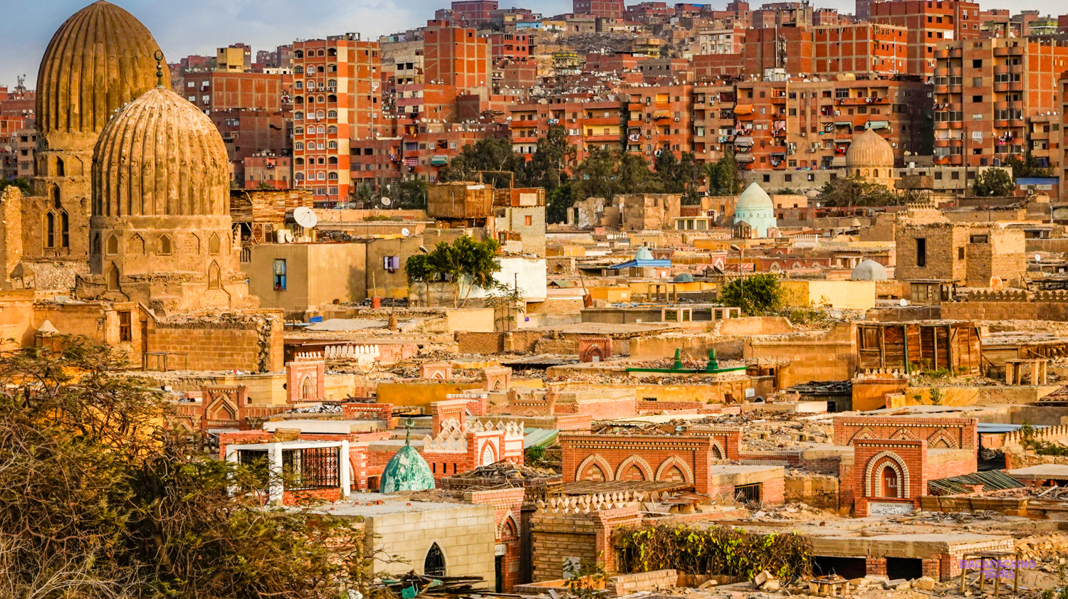 Ancient domed tombs and historical buildings fill the sprawling city of Cairo under a cloudy sky.