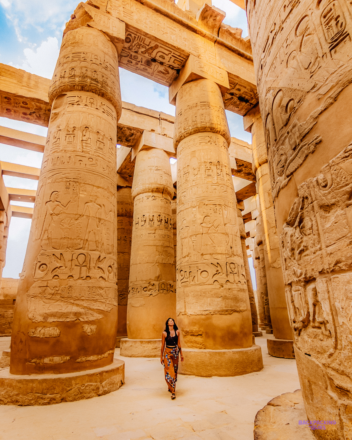 A woman explores the grand, hieroglyph-covered columns of Karnak Temple under a bright blue sky.