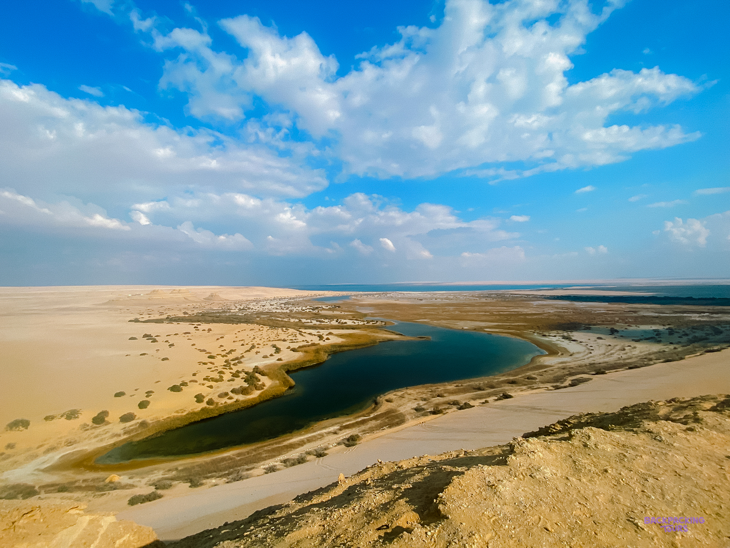 A serene desert landscape in Faiyum, Egypt, with a winding blue lake under a vast sky.