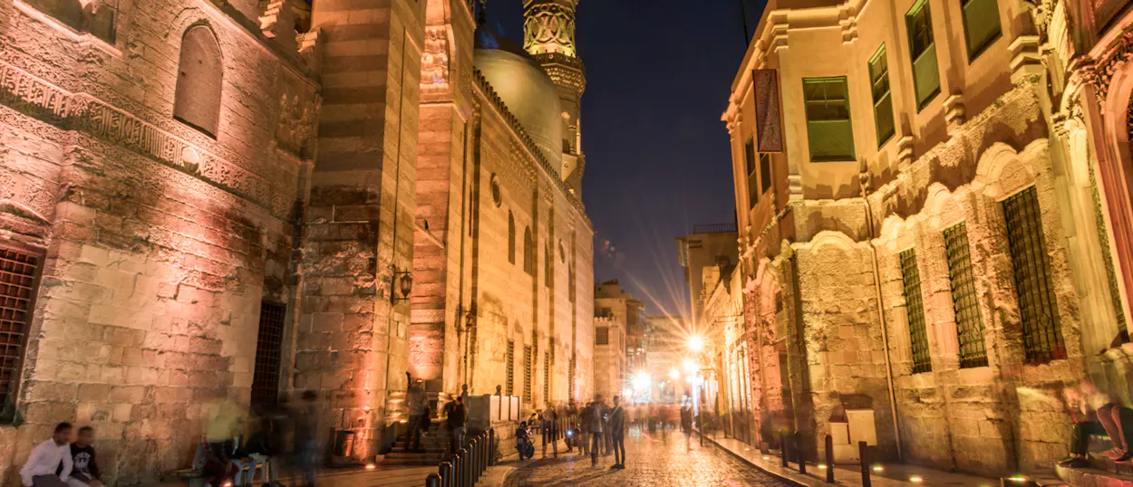 Warm lights illuminate a historic street in Cairo at night, with blurred people and ornate architecture.