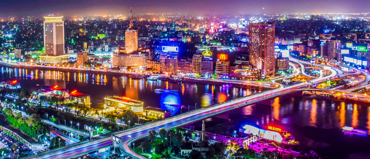 Vibrant Cairo cityscape at night with illuminated buildings, bridges, and light trails over the Nile River.