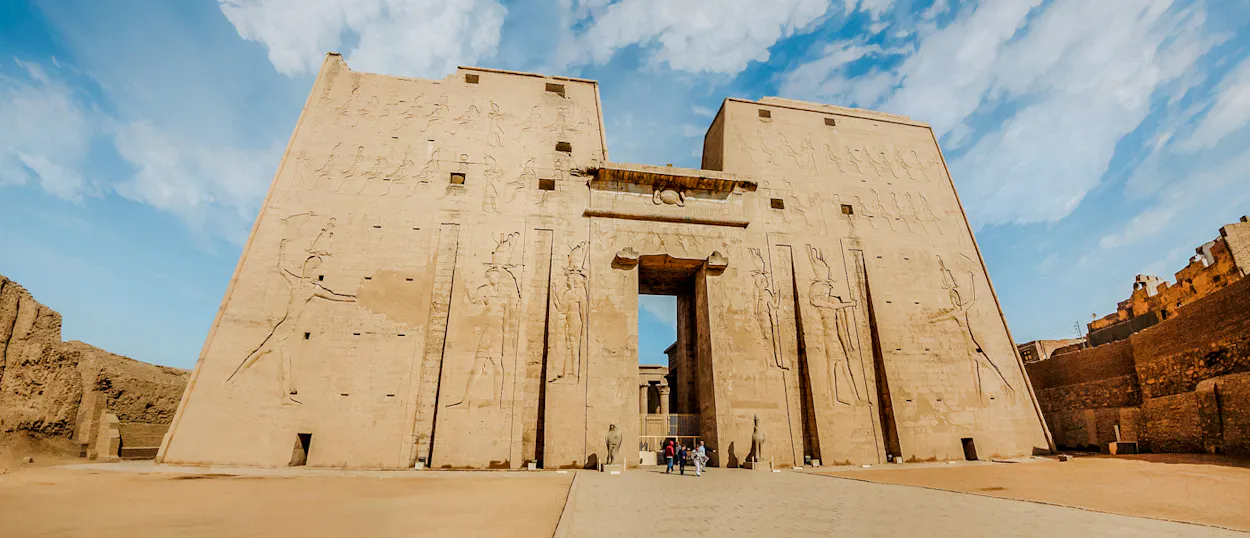 Tourists admire the towering ancient Edfu Temple entrance with detailed carved reliefs under a bright sky.