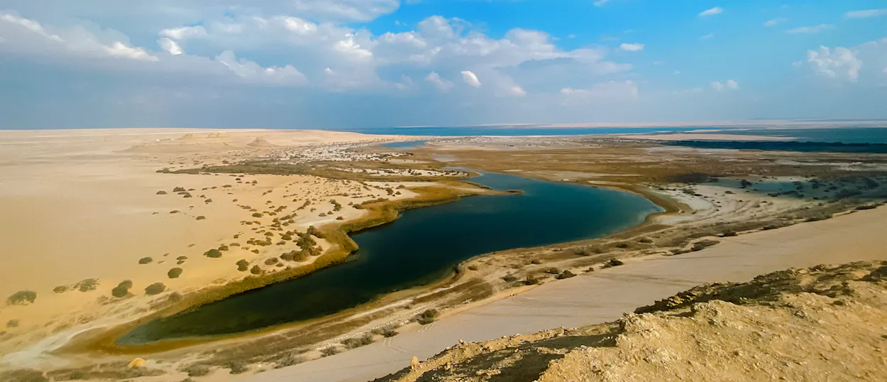 A dark blue desert lake winds through golden sands under a bright, partly cloudy sky in Faiyum.