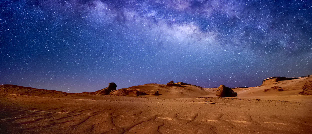 The Milky Way stretches across a dark desert sky above ancient rock formations in Wadi al Hitan.