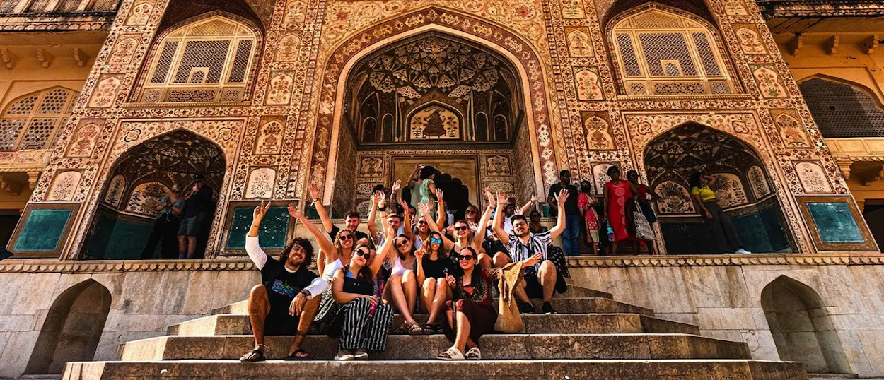 A group of travellers sits on stone steps, smiling and waving in front of an ornately decorated building.