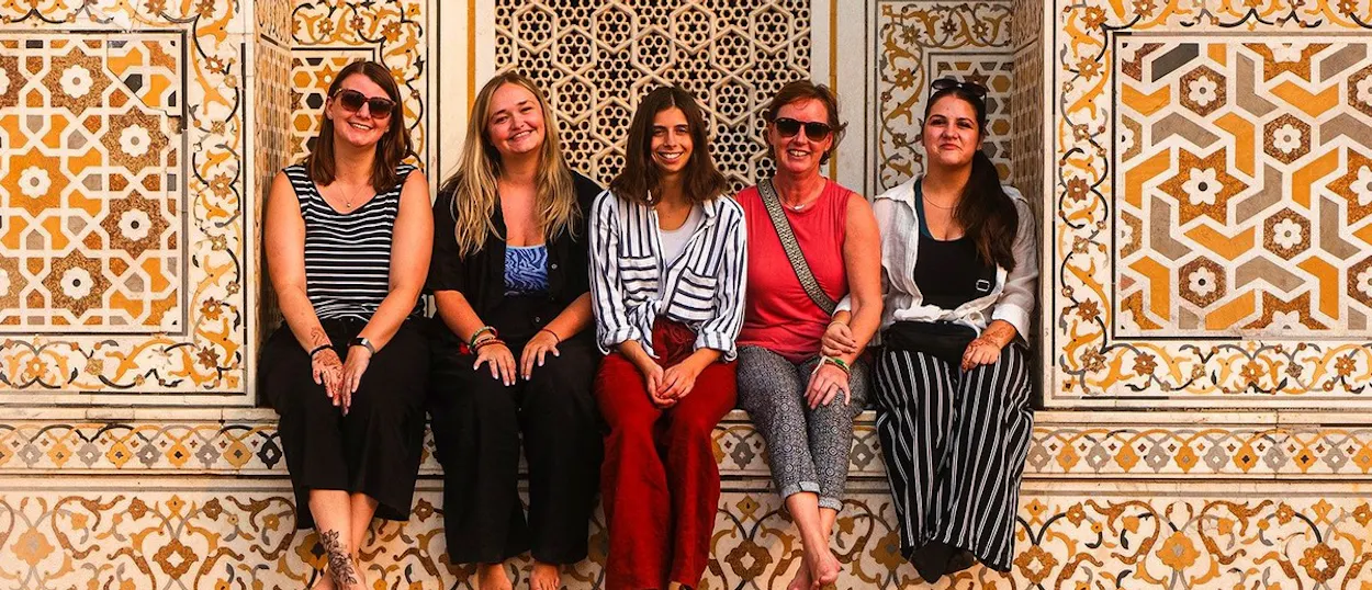 Five women sit barefoot against an ornate wall decorated with yellow and grey geometric patterns.