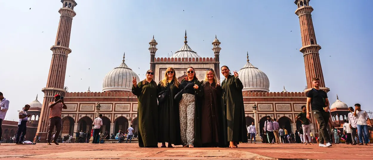 Five women pose on a large red courtyard in front of a grand mosque with domes and minarets.