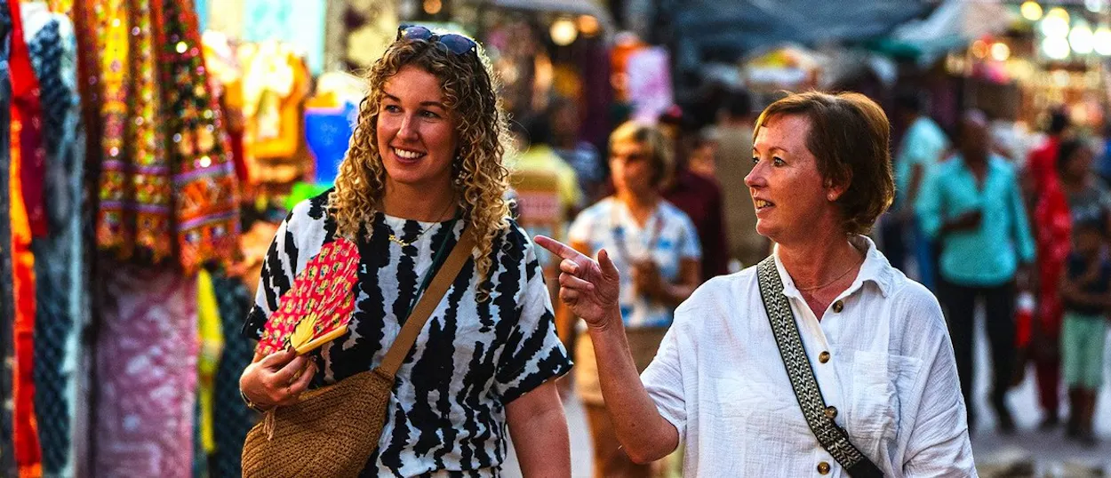 Two women walk through a bustling market street, one smiling and holding a red fan.