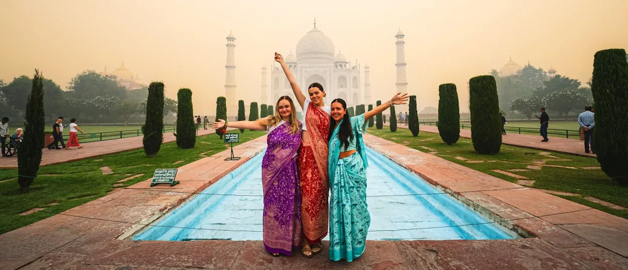 Three women in colourful saris pose with outstretched arms before a majestic white domed building and reflecting pool.