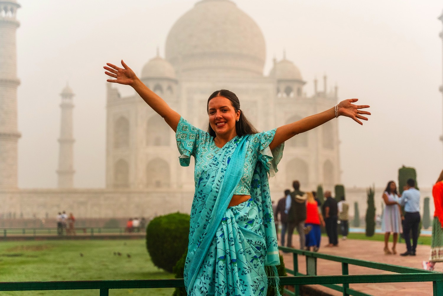 A traveller in a turquoise sari throws her arms wide in front of the Taj Mahal on a misty morning.