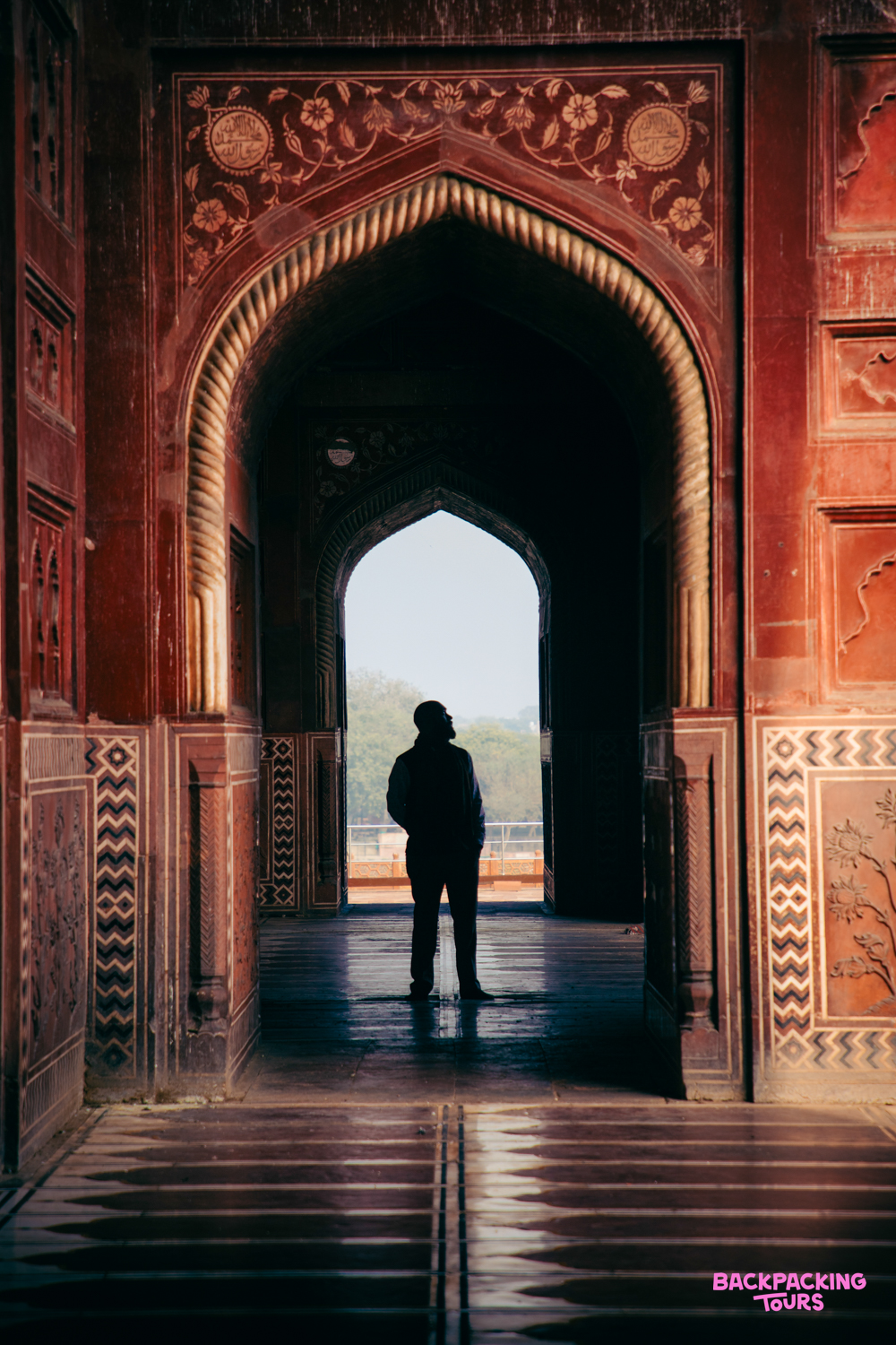 A person stands silhouetted in an ornate red archway, gazing out at the hazy view in Agra.