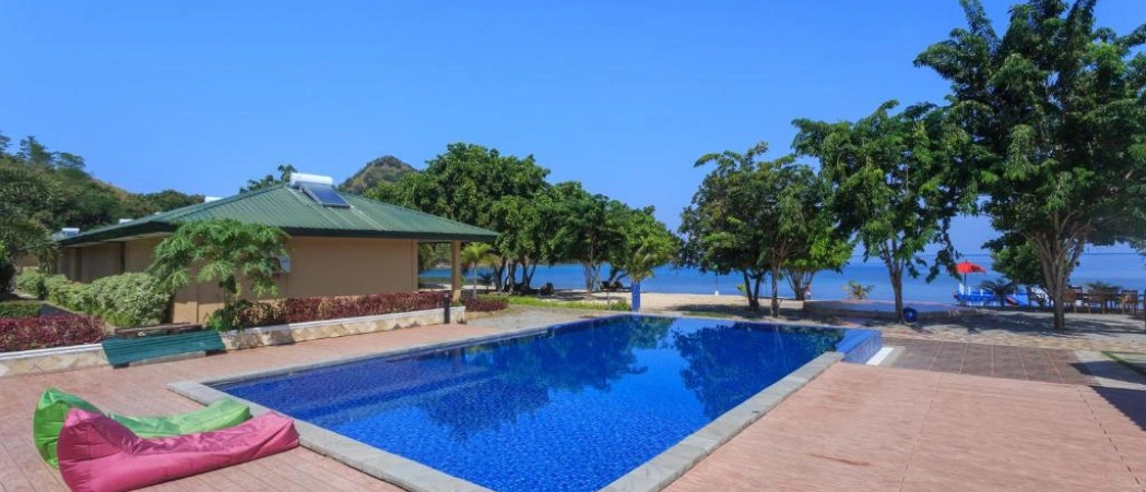 A tropical resort in Labuan Bajo featuring a blue swimming pool, lush trees, and a sandy beach.