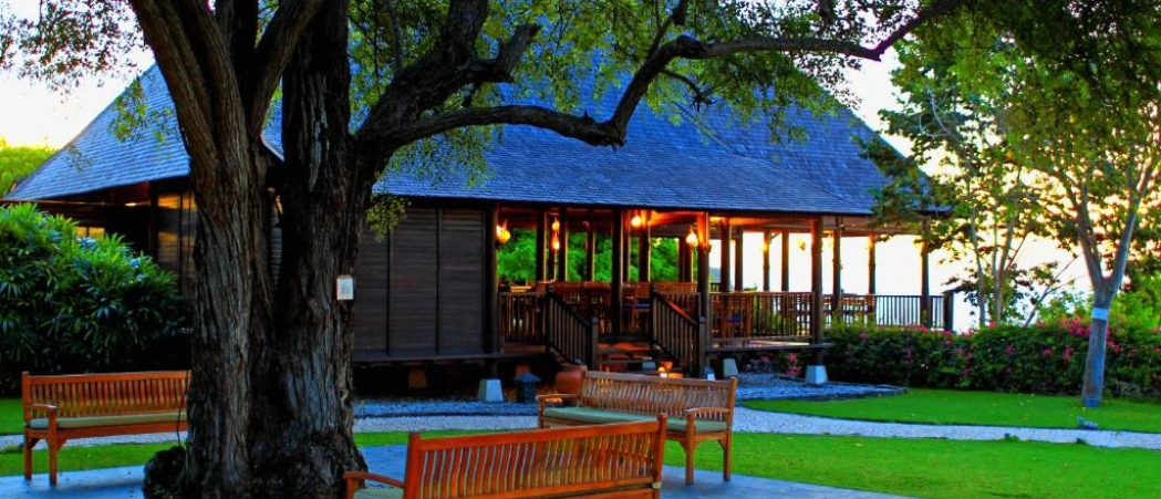 A traditional guesthouse with a dark roof and illuminated porch, surrounded by green grass and trees in Sumbawa.