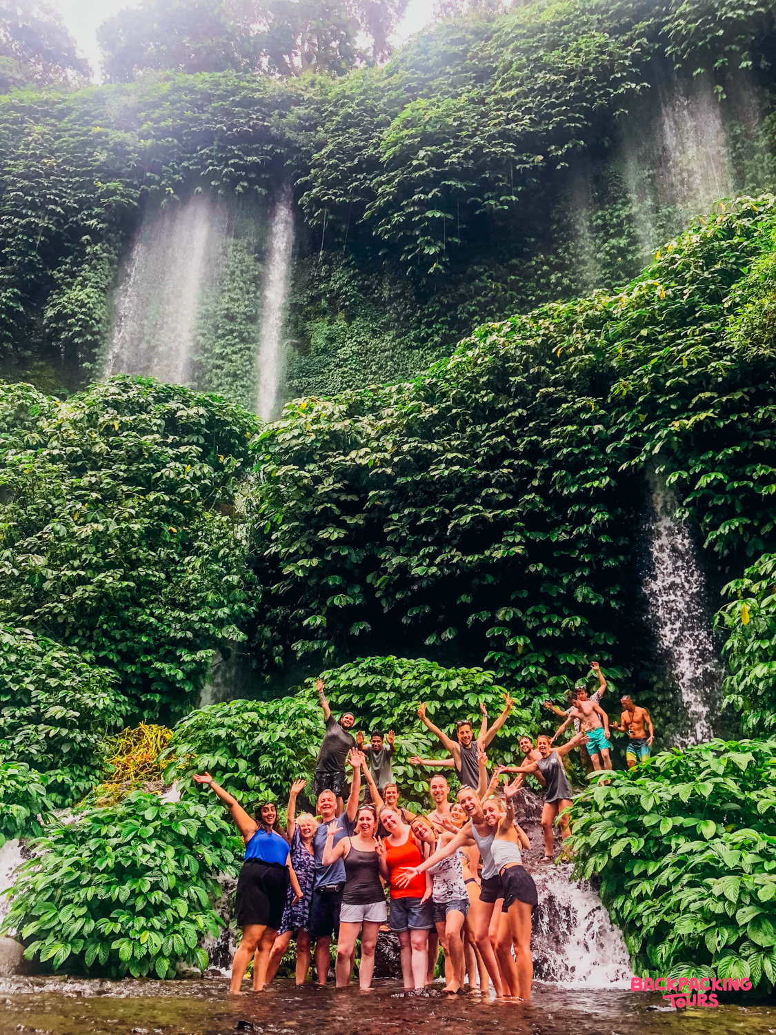 Excited travelers pose in shallow water before multiple waterfalls cascading down a lush green cliff in Lombok.