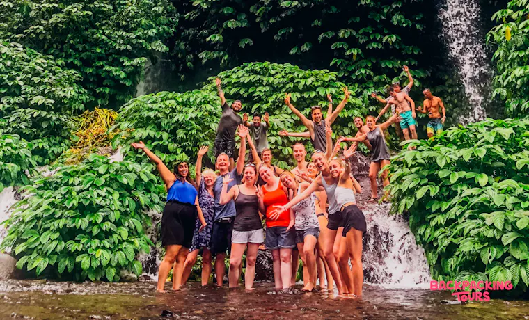 Excited travelers pose in shallow water before multiple waterfalls cascading down a lush green cliff in Lombok.