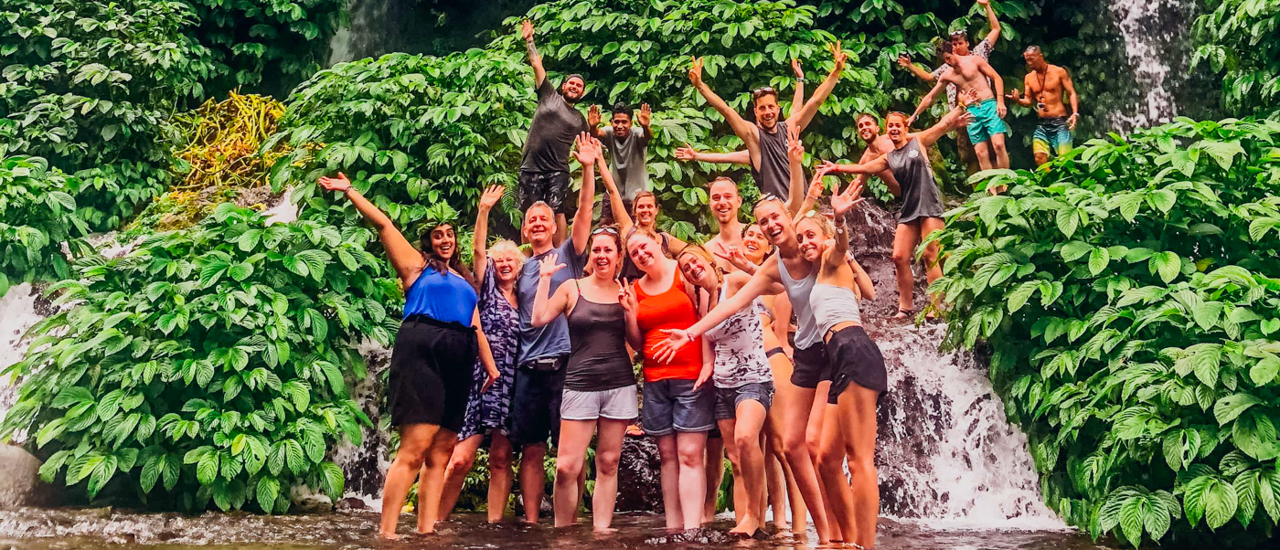 A large group of happy travelers pose in front of lush green waterfalls in Lombok, Indonesia.