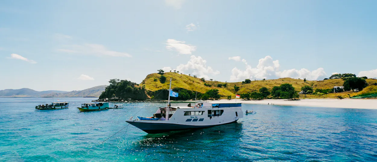 A white live-aboard boat floats on clear turquoise water near Satonda Island's sunny beach.