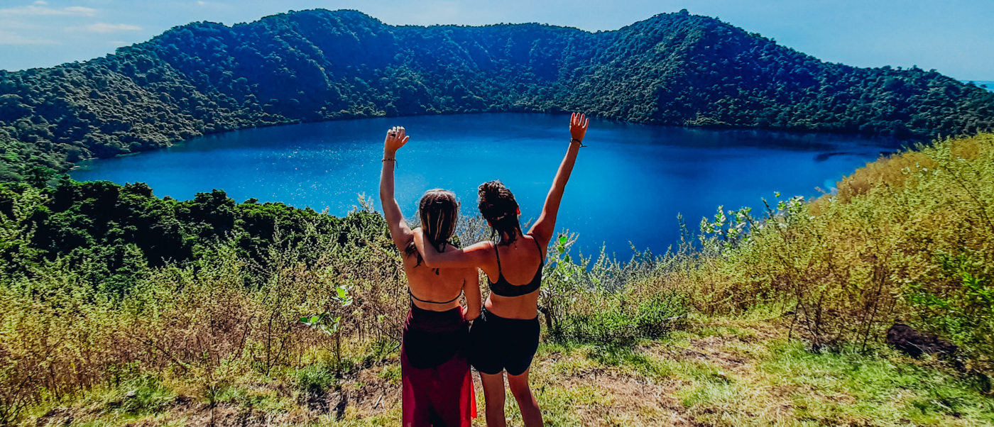 Two women with raised arms celebrate the stunning view of Satonda Island's blue crater lake.