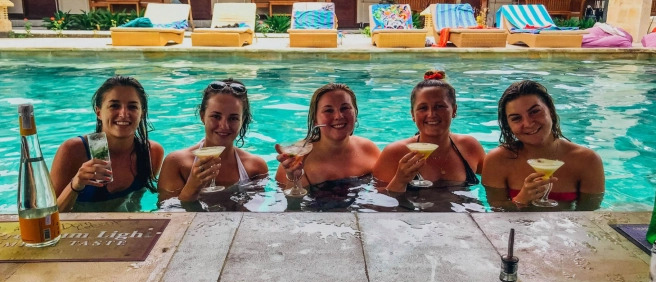 Five happy women smiling with cocktails at a swim-up bar in a pool in Lombok.