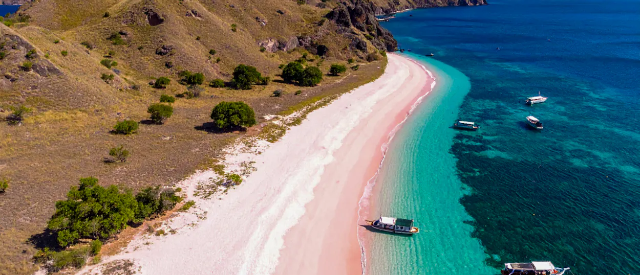 Pink Beach in Komodo National Park, with clear turquoise water, anchored boats, and dry hills.
