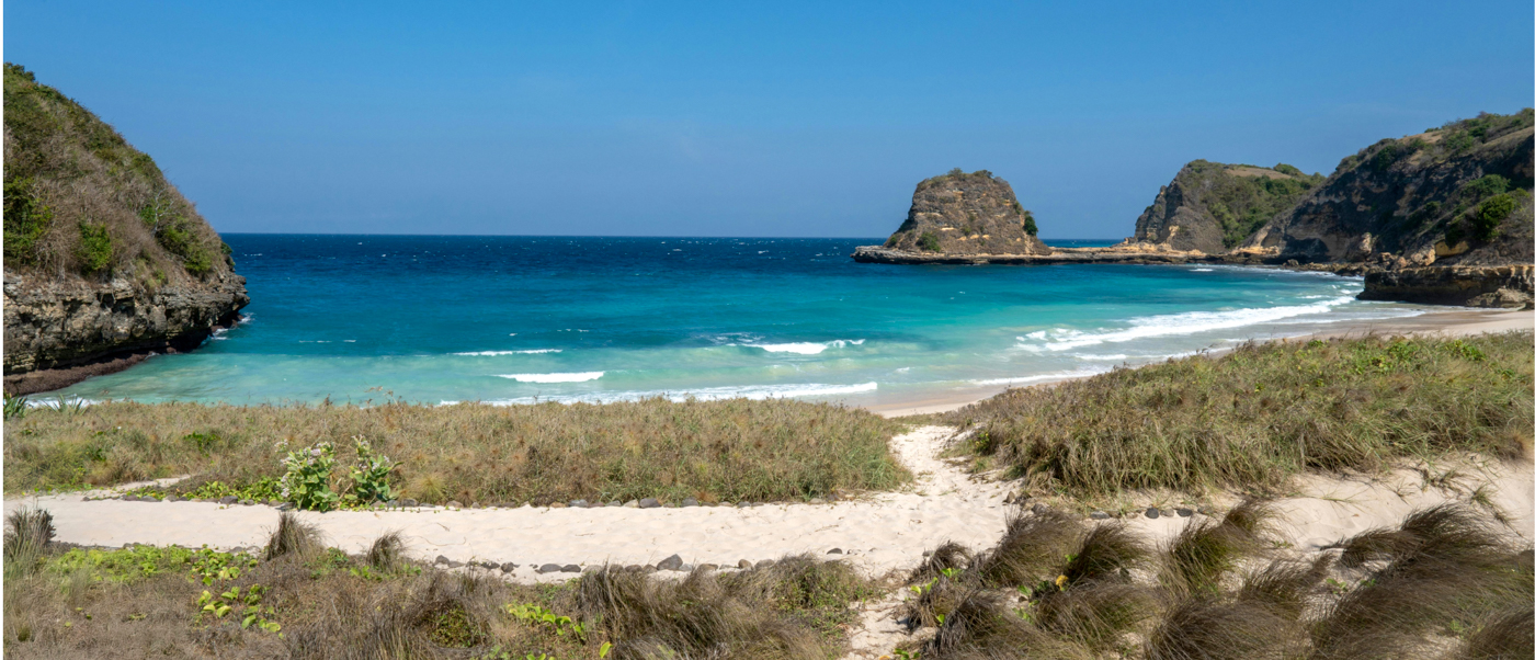 Turquoise ocean water laps a sandy beach with rocky cliffs and dry grass on Lombok.