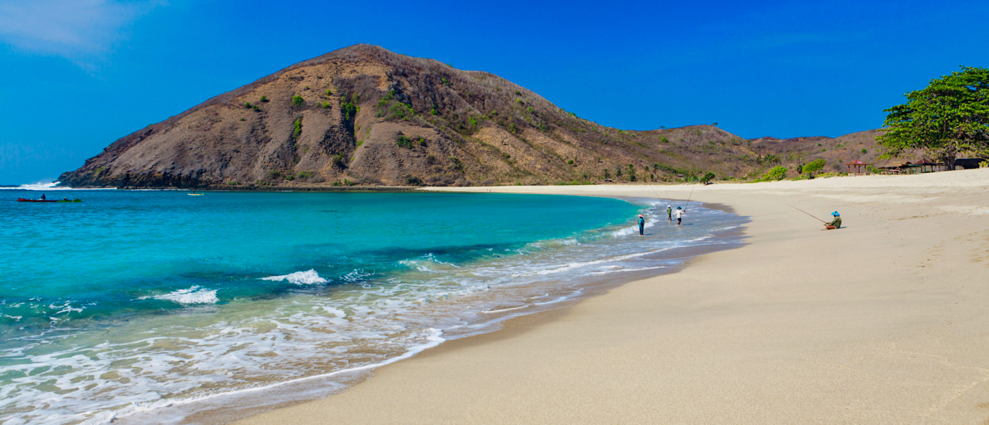 Beautiful white sand beach with turquoise water, gentle waves, and a large dry mountain in Lombok.