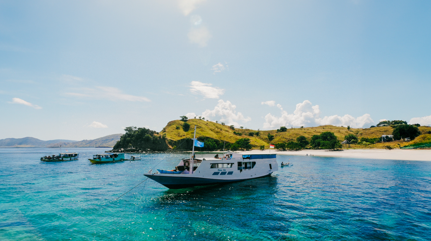 A white boat floats on clear turquoise water near a white sand beach and green hills in Komodo.