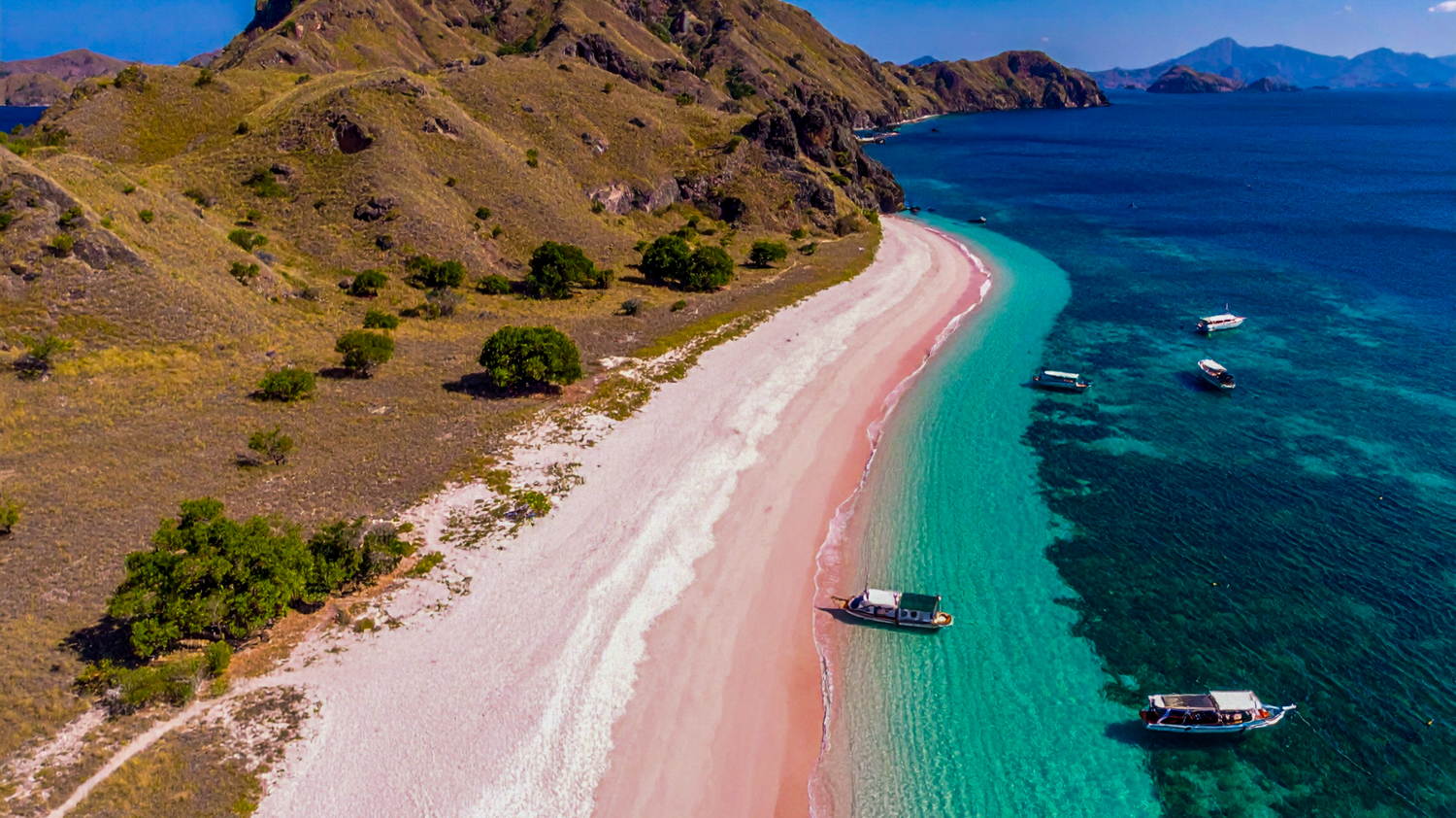 Aerial view of Pink Beach, Komodo Island, with clear turquoise water and several boats anchored.