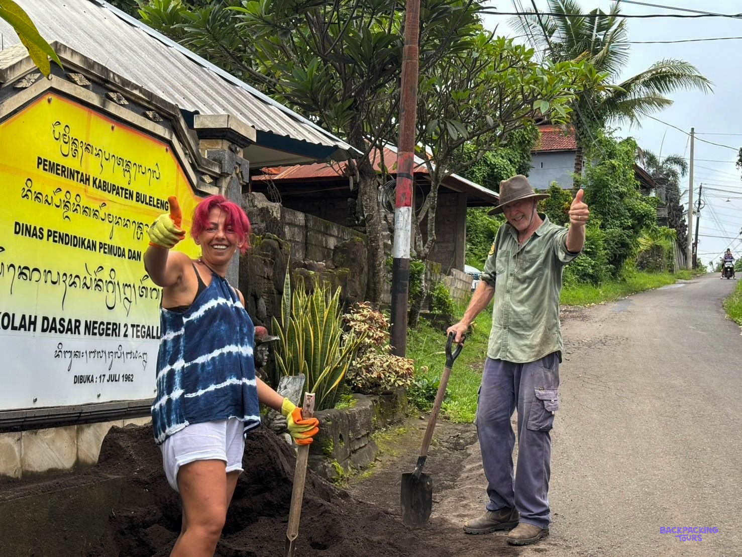 Two volunteers with spades give a thumbs-up beside the sign of a local primary school.