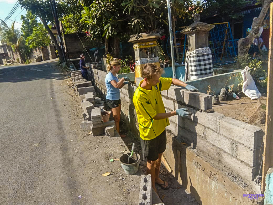 A volunteer in a yellow shirt lays concrete blocks for a new wall beside a village lane.