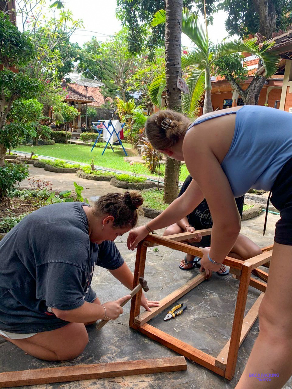 Two volunteers hammer together the wooden frame of a table in a leafy garden.