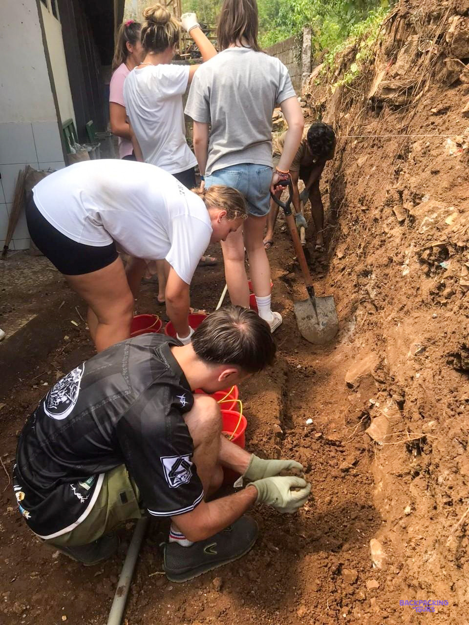 Volunteers dig out a trench of red earth beside a school building with buckets and trowels.