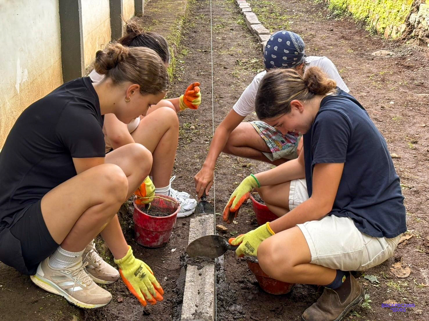 Three volunteers crouch with buckets of cement, setting stones along the edge of a school path.