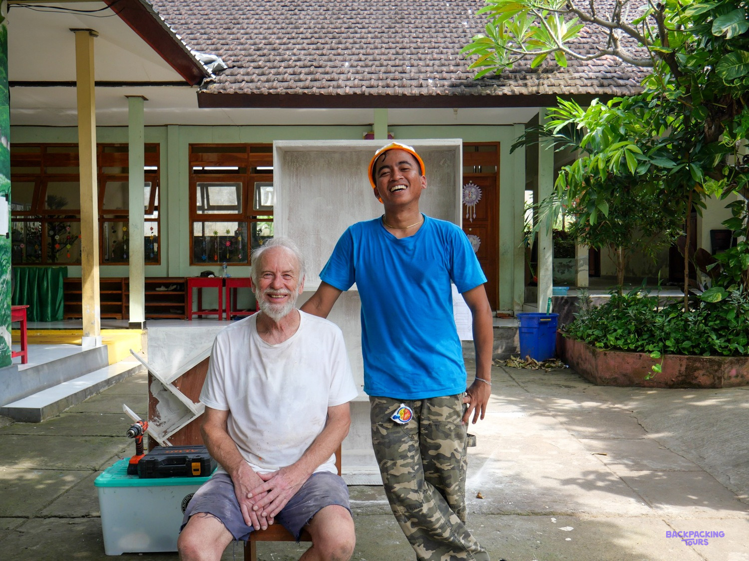 A local builder in an orange cap laughs beside a volunteer resting on a toolbox at a school.