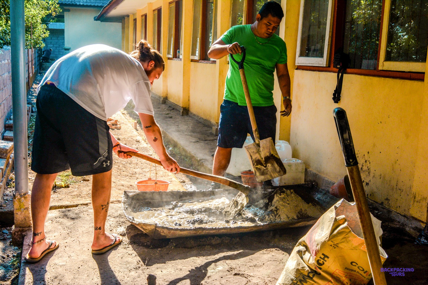 A volunteer mixes cement in a tray beside a school wall as a local builder digs alongside.