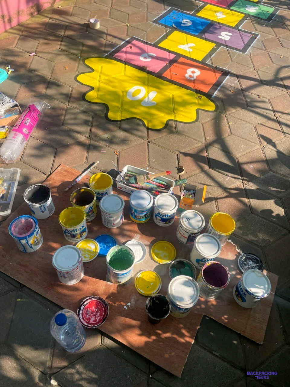 Dozens of paint pots sit on a board beside a freshly painted hopscotch grid.