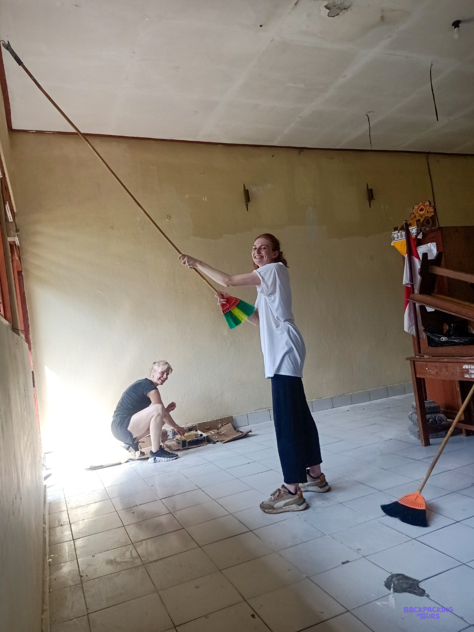 A volunteer paints a classroom wall with a long roller while another works on the floor.